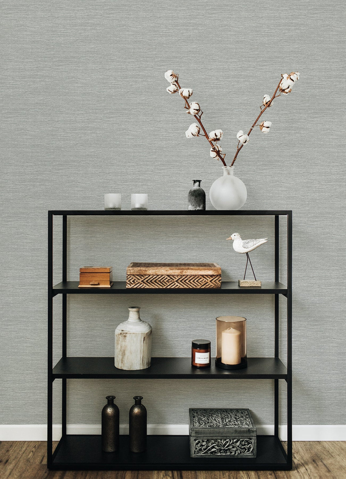 A black metal shelf holds decorative items—vases, a wooden box, candles, a bird figurine, two bottles, and a patterned box—set against walls covered in Malin Faux Grasscloth Wallpaper by York Wallcoverings on a wooden floor.