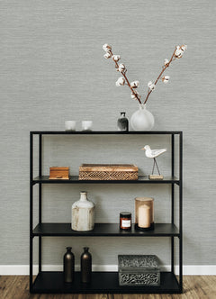 A black metal shelf holds decorative items—vases, a wooden box, candles, a bird figurine, two bottles, and a patterned box—set against walls covered in Malin Faux Grasscloth Wallpaper by York Wallcoverings on a wooden floor.