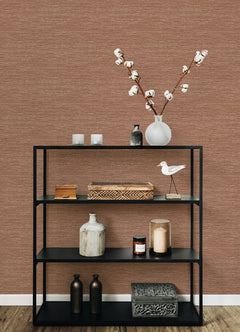 A black metal shelf styled with decorative items—including vases, candles, boxes, and a white ceramic bird—sits in front of a wall featuring Malin Faux Grasscloth Wallpaper by York Wallcoverings. On the top shelf, a glass vase filled with cotton stems stands out against the wallpaper’s rich brown texture.