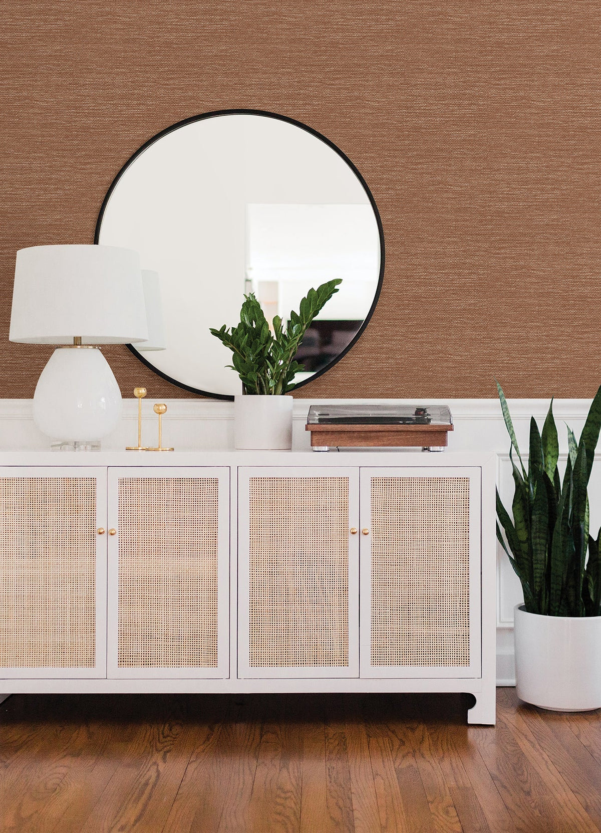 A white sideboard with rattan doors holds a lamp, plant, and decor items. A round mirror hangs above, and a tall potted plant stands to the right. The wall features York Wallcoverings’ Malin Faux Grasscloth Wallpaper in brown, complementing the wooden floor.