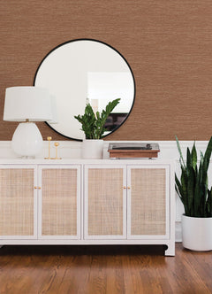 A white sideboard with rattan doors holds a lamp, plant, and decor items. A round mirror hangs above, and a tall potted plant stands to the right. The wall features York Wallcoverings’ Malin Faux Grasscloth Wallpaper in brown, complementing the wooden floor.