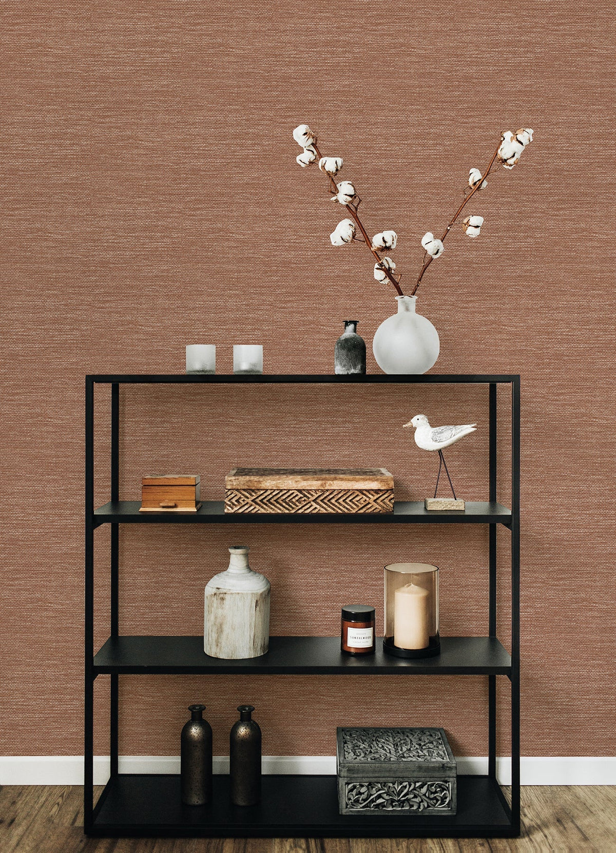 A black metal shelf adorned with decorative items—including vases, candles, boxes, and a cotton branch in a white vase—stands against a wall covered in York Wallcoverings’ Malin Faux Grasscloth Wallpaper, with a wooden floor beneath.