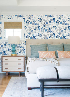 A bright bedroom featuring York Wallcoverings’ Jonah Leaf Trail Wallpaper, a beige tufted bed adorned with blue and peach pillows, a white bench, a blue lamp on a wooden nightstand, and a window dressed with a bamboo shade.