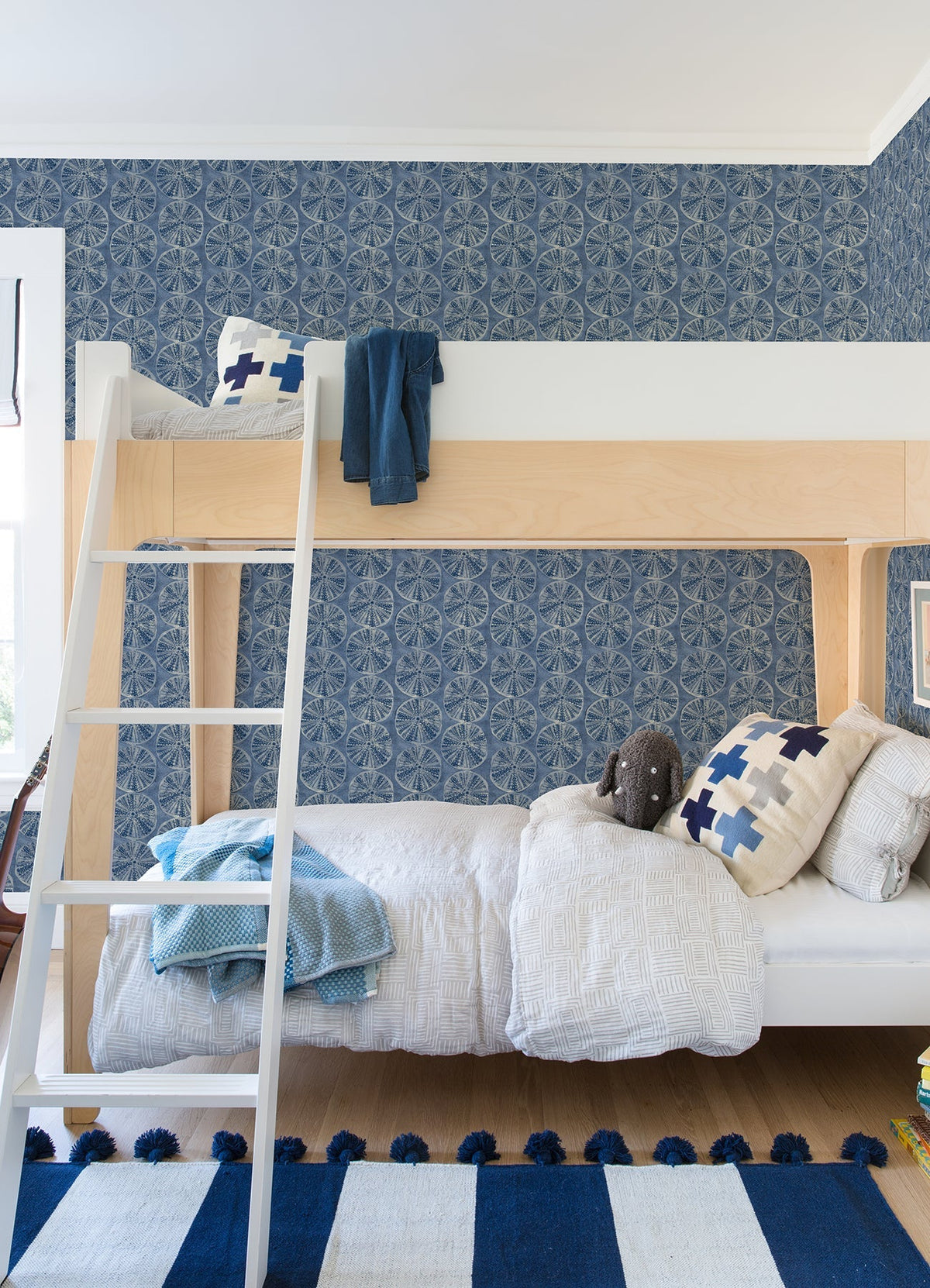Modern kids’ bedroom featuring a wooden bunk bed, blue and white bedding, Chesapeake Sea Biscuit Sand Dollar Wallpaper by York Wallcoverings, a plush toy, a ladder, and a striped blue and white rug on a wooden floor.