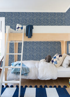 Modern kids’ bedroom featuring a wooden bunk bed, blue and white bedding, Chesapeake Sea Biscuit Sand Dollar Wallpaper by York Wallcoverings, a plush toy, a ladder, and a striped blue and white rug on a wooden floor.