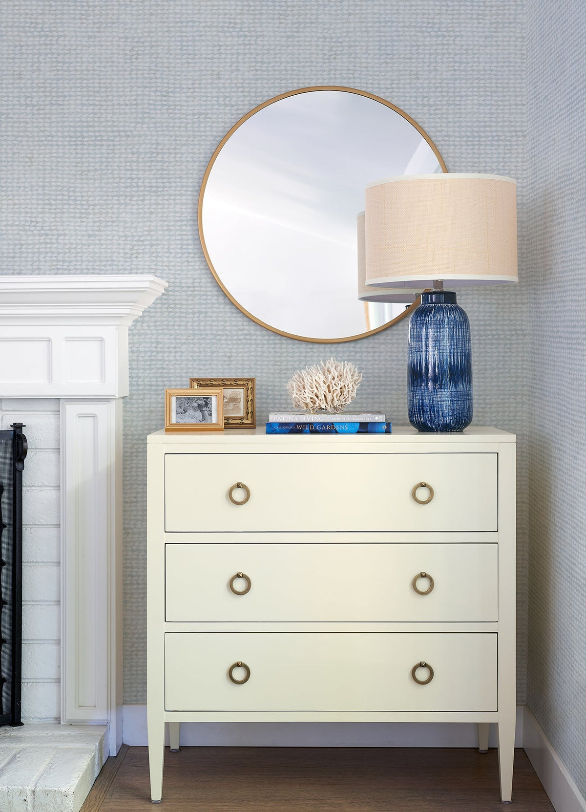 A cream dresser with three drawers sits beside a white fireplace, set against Chesapeake Wellen Abstract Rope Wallpaper by York Wallcoverings. On top are a blue lamp, a white coral, books, and framed photos. A round mirror hangs above, reflecting part of the room.