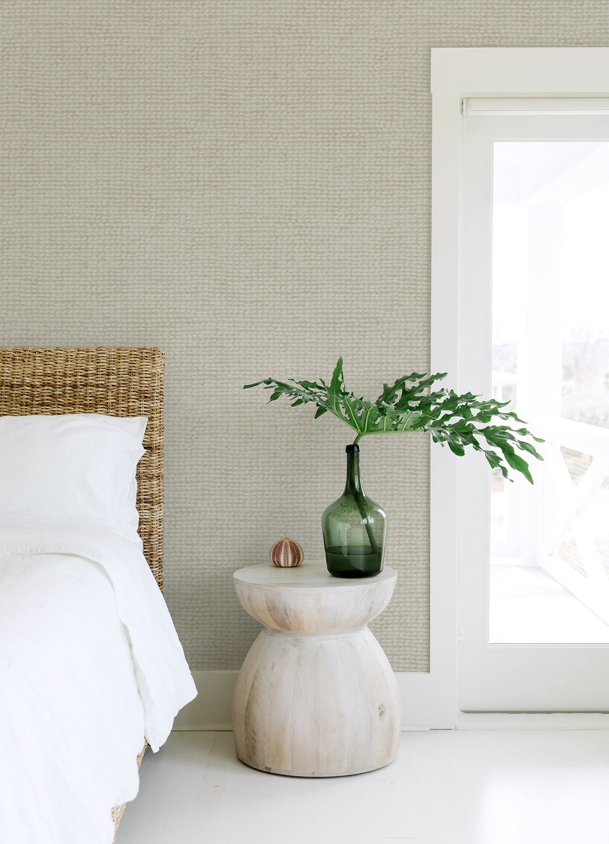 A minimalist bedroom featuring Chesapeake Wellen Abstract Rope Wallpaper by York Wallcoverings, a wicker bed with white bedding, a small round wooden side table, a green glass vase filled with leafy branches, and a white-framed glass door allowing natural light to stream in.