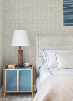 A modern bedroom featuring a beige upholstered bed with white bedding and blue-accented pillows, complemented by a nightstand displaying a wooden lamp, box, glasses, and mug. The light-colored wall is adorned with York Wallcoverings’ Chesapeake Wellen Abstract Rope Wallpaper.