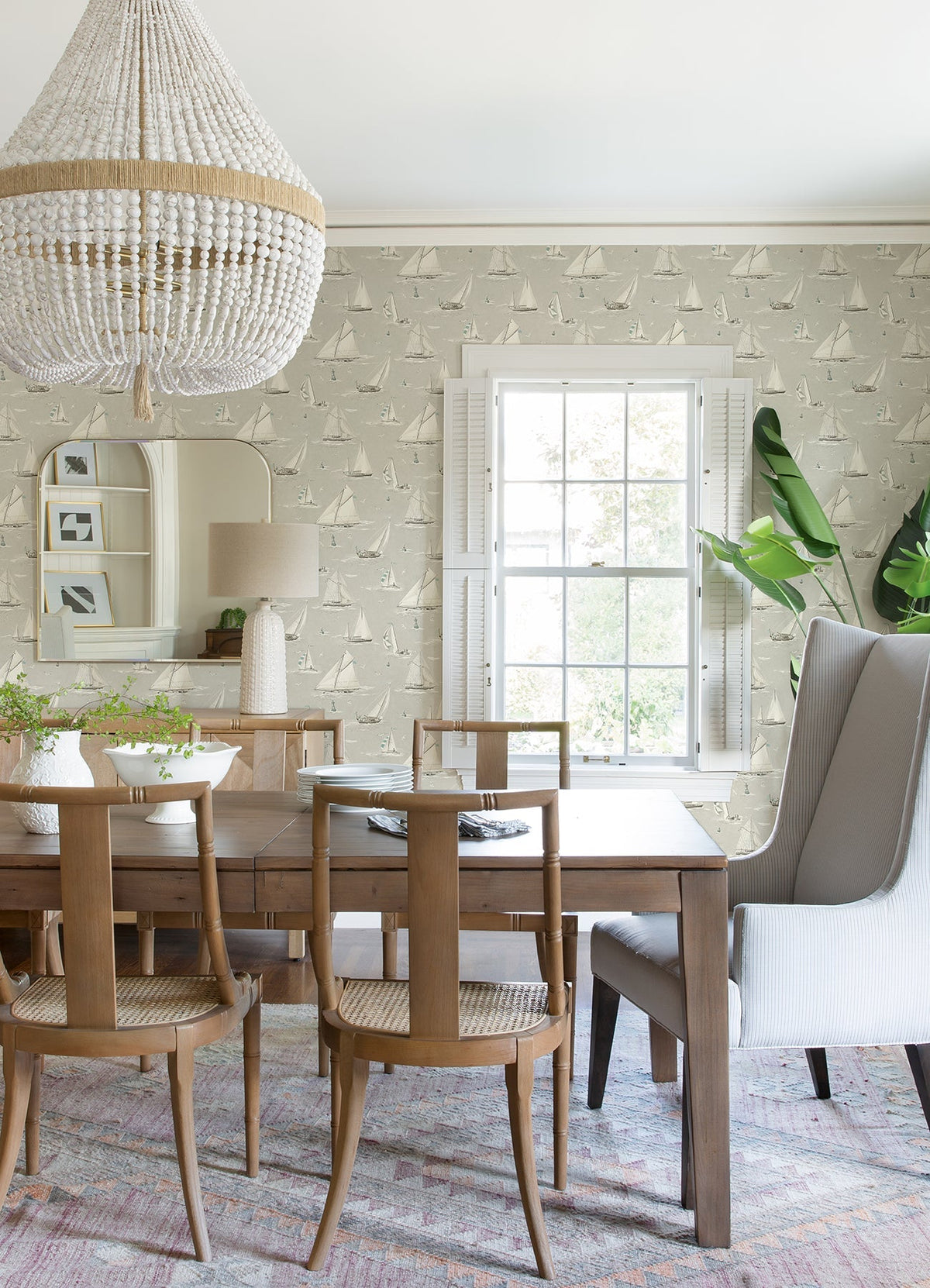 Bright dining room with a wood table, six chairs, a large beaded chandelier, tall plant, a window with white shutters, pastel rug on the floor, and walls adorned with Chesapeake Leeward Sailboat Wallpaper by York Wallcoverings.