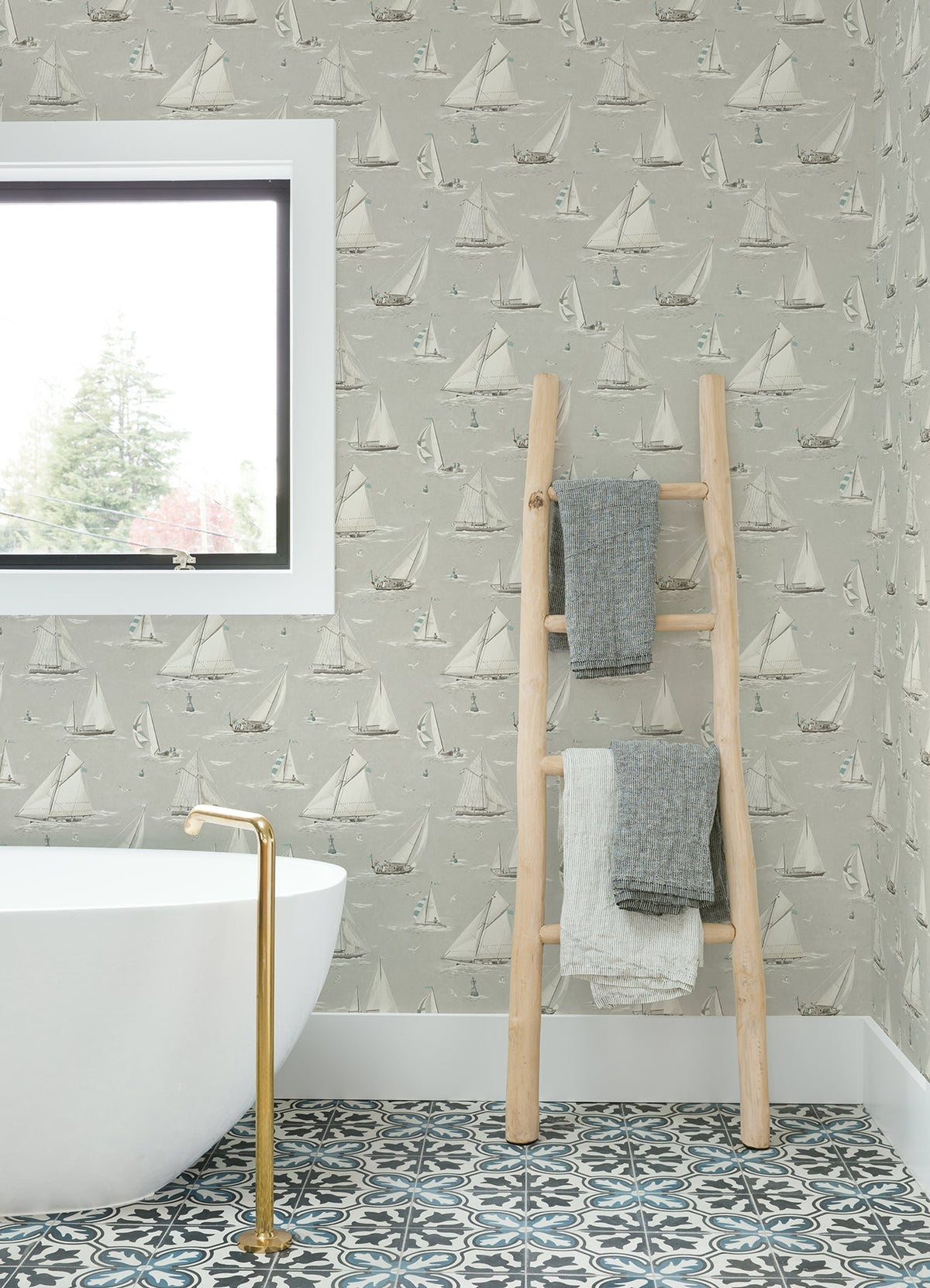A bathroom featuring a white tub, gold faucet, patterned tile floor, window, and a wooden ladder holding three folded towels is adorned with York Wallcoverings’ Chesapeake Leeward Sailboat Wallpaper.