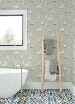 A bathroom featuring a white tub, gold faucet, patterned tile floor, window, and a wooden ladder holding three folded towels is adorned with York Wallcoverings’ Chesapeake Leeward Sailboat Wallpaper.