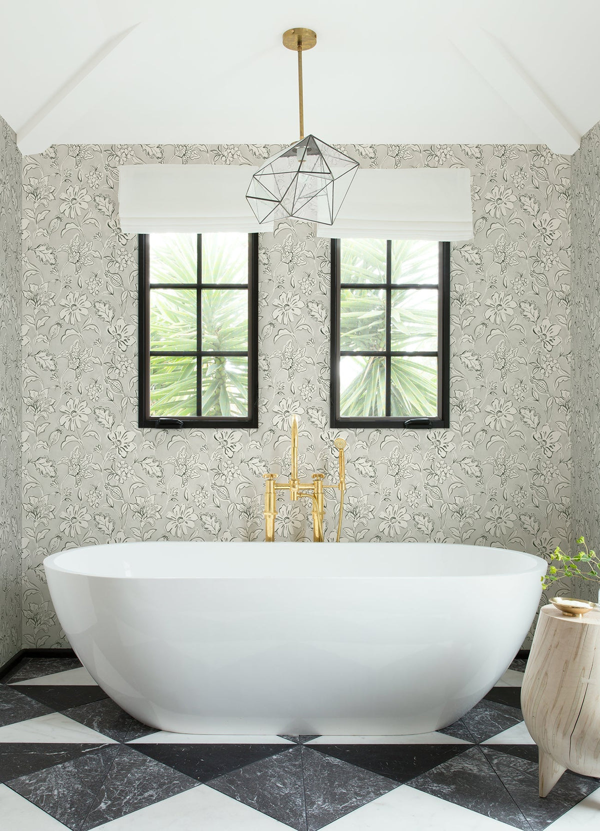 Modern bathroom with a white oval bathtub, gold faucet, geometric pendant light, two windows, black-and-white checkered floor tiles, and York Wallcoverings Chesapeake Plumeria Floral Trail Wallpaper.