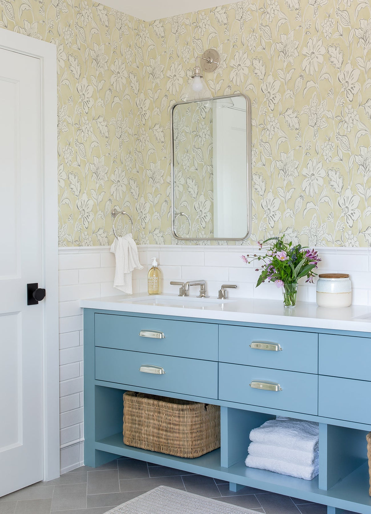 A bathroom featuring a blue vanity with double sinks, a rectangular mirror, white tiled walls adorned with York Wallcoverings Chesapeake Plumeria Floral Trail Wallpaper, a basket, towels, a vase of flowers, and soap on the counter.