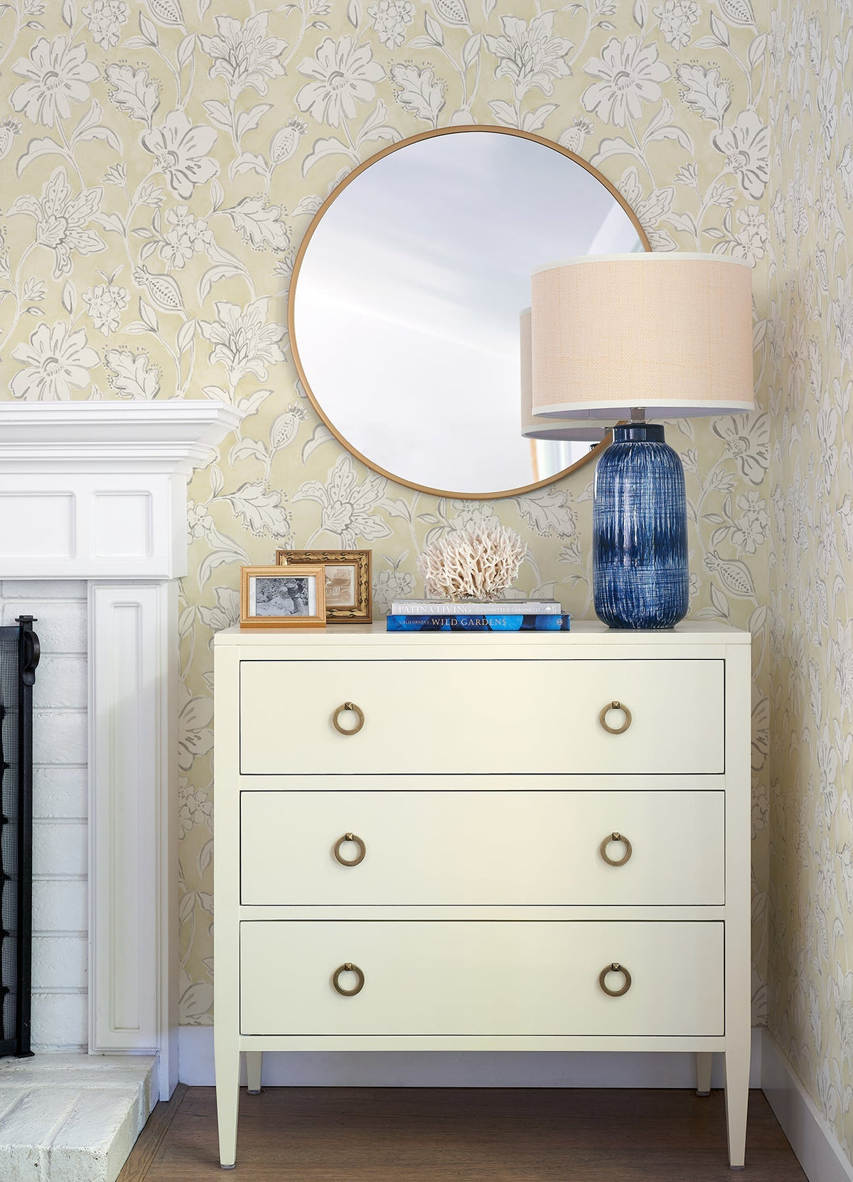 A white dresser with three drawers sits below a round mirror. On top are a blue lamp with a beige shade, books, coral decor, and framed photos. The background features Chesapeake Plumeria Floral Trail Wallpaper by York Wallcoverings and part of a white fireplace.