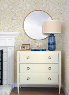 A white dresser with three drawers sits below a round mirror. On top are a blue lamp with a beige shade, books, coral decor, and framed photos. The background features Chesapeake Plumeria Floral Trail Wallpaper by York Wallcoverings and part of a white fireplace.