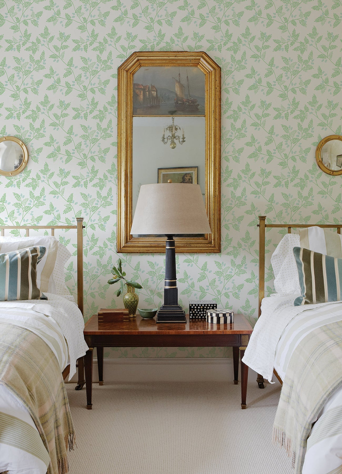 A bedroom with two twin beds, a wooden nightstand between them, a table lamp, a gold-framed mirror on the walls covered in York Wallcoverings’ Ardell Botanical Wallpaper, and striped pillows and throws on each bed.
