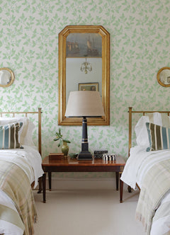A bedroom with two twin beds, a wooden nightstand between them, a table lamp, a gold-framed mirror on the walls covered in York Wallcoverings’ Ardell Botanical Wallpaper, and striped pillows and throws on each bed.