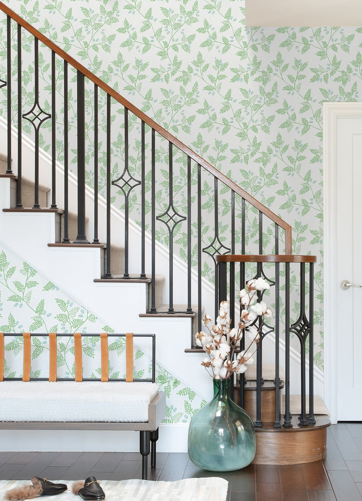 A staircase with black iron railings, Ardell Botanical Wallpaper by York Wallcoverings featuring a green leaf pattern, a white bench with leather straps, a large green vase holding cotton stems, and dark wood floors.