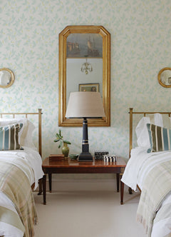 Two twin beds with striped pillows and light plaid throws sit on either side of a wooden nightstand topped with a lamp, books, and a plant. A large mirror and framed art hang on a wall adorned with Ardell Botanical Wallpaper by York Wallcoverings.