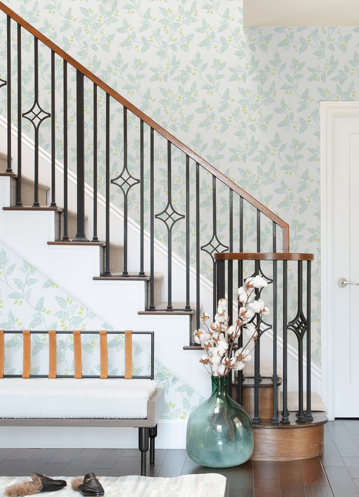 A staircase with black iron rails and a wooden handrail stands next to a bench with leather straps. Against the wall, covered in Ardell Botanical Wallpaper by York Wallcoverings, a large green vase filled with cotton stems sits on dark wood flooring.