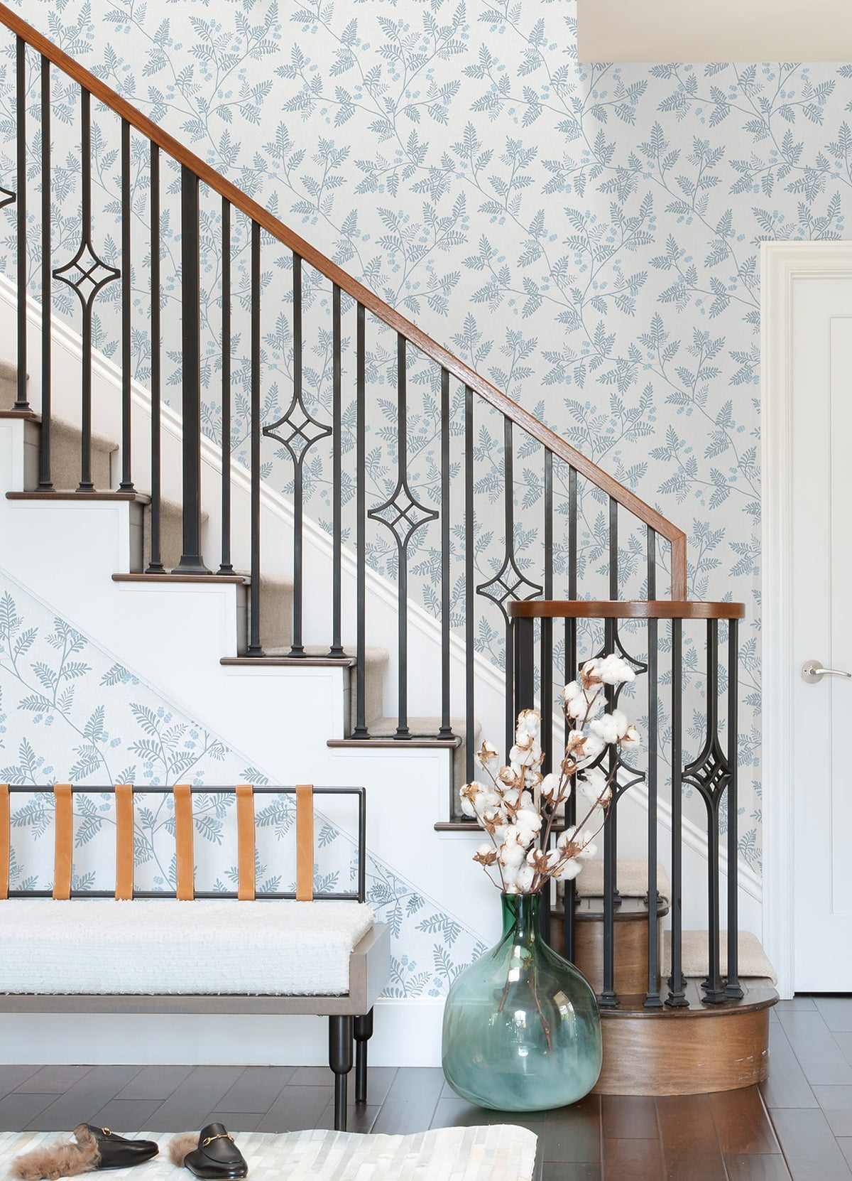 A staircase with a black metal railing and wood handrail, Ardell Botanical Wallpaper by York Wallcoverings, a bench with leather straps, a large green vase with cotton stems, and brown shoes on a dark wood floor.