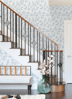 A staircase with a black metal railing and wood handrail, Ardell Botanical Wallpaper by York Wallcoverings, a bench with leather straps, a large green vase with cotton stems, and brown shoes on a dark wood floor.