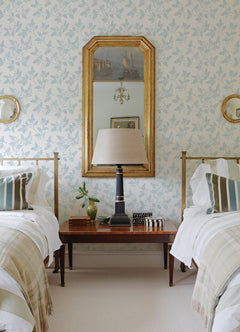 Two twin beds with striped pillows and plaid blankets flank a wooden table holding a lamp, books, and a plant. The wall is decorated with Ardell Botanical Wallpaper by York Wallcoverings, complemented by a gold-framed mirror and round mirrors on each side.