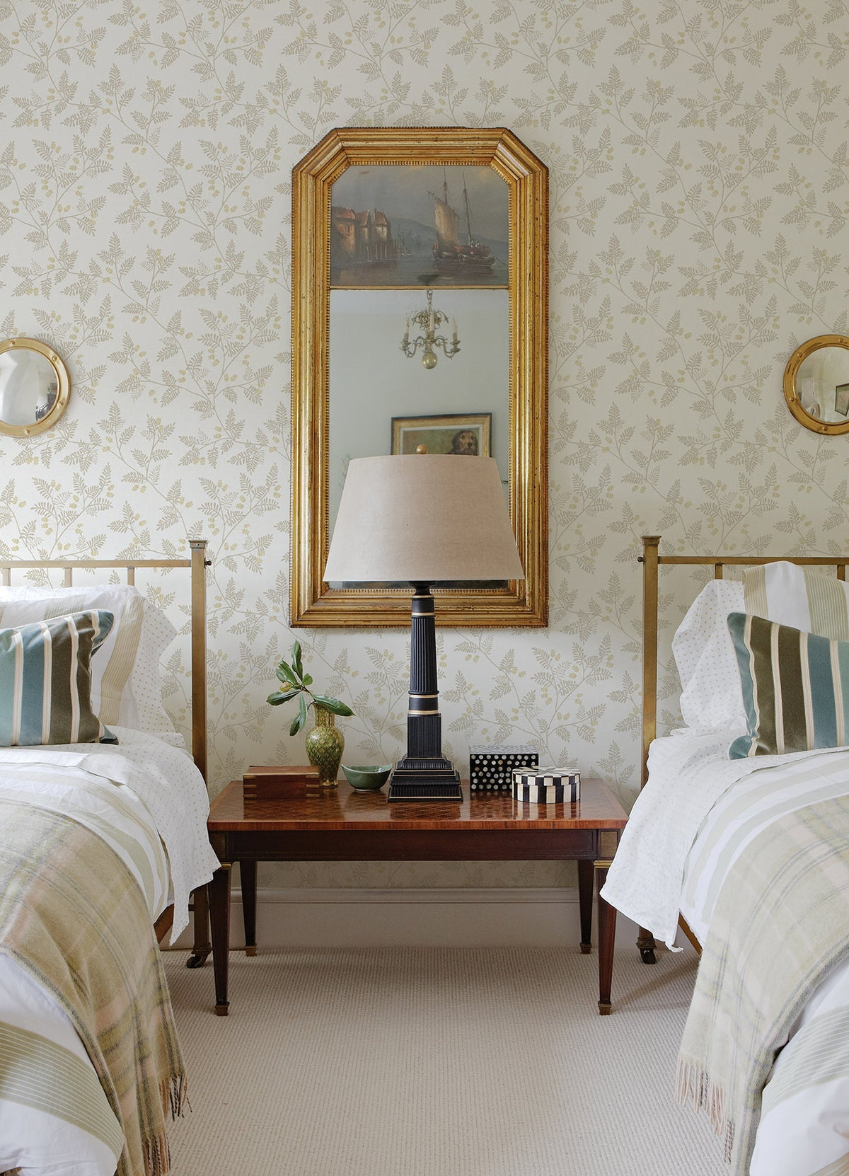 Two single beds with striped pillows flank a wooden nightstand holding a lamp, small plant, and decor; a gold-framed mirror hangs on the wall behind, which is adorned with Ardell Botanical Wallpaper by York Wallcoverings.
