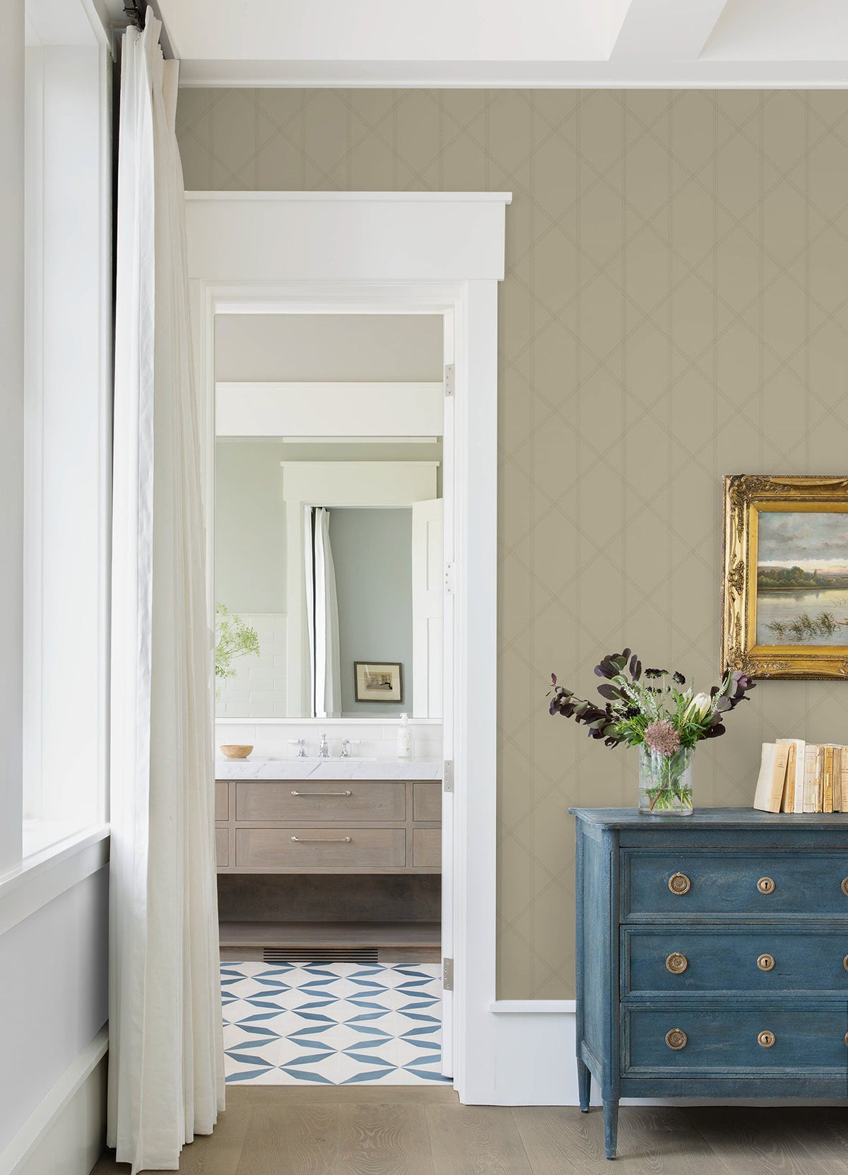 A room featuring Walcott Stitched Trellis Wallpaper by York Wallcoverings on the walls, a blue dresser topped with a vase of flowers, a gold-framed painting, and a doorway opening to a bathroom with a wood vanity and geometric tile floor. White curtains hang beside the window.