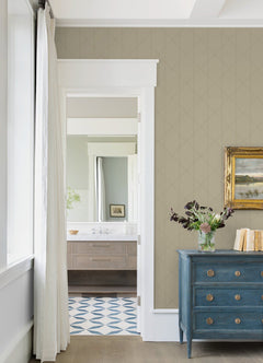 A room featuring Walcott Stitched Trellis Wallpaper by York Wallcoverings on the walls, a blue dresser topped with a vase of flowers, a gold-framed painting, and a doorway opening to a bathroom with a wood vanity and geometric tile floor. White curtains hang beside the window.