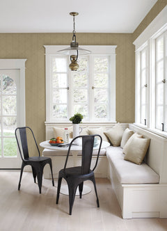 A bright breakfast nook featuring York Wallcoverings’ Walcott Stitched Trellis Wallpaper, a white round table, two black metal chairs, a built-in white bench with beige cushions, and large windows flooding the space with natural light. A fruit bowl and plant adorn the table.