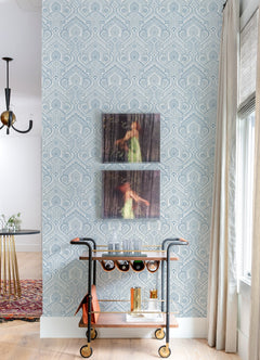 A bar cart adorned with bottles and glasses stands against a wall covered in Fernback Ornate Botanical Wallpaper by York Wallcoverings. Above the cart, two blurred art pieces feature a person in a green dress. To the right, there is a window with a curtain, while part of the dining room can be seen to the left.