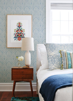 A mid-century nightstand with a lamp, a small vase of flowers, and framed art above sits next to a bed with blue and white pillows and a dark blue throw in a bright bedroom featuring Fernback Ornate Botanical Wallpaper by York Wallcoverings.