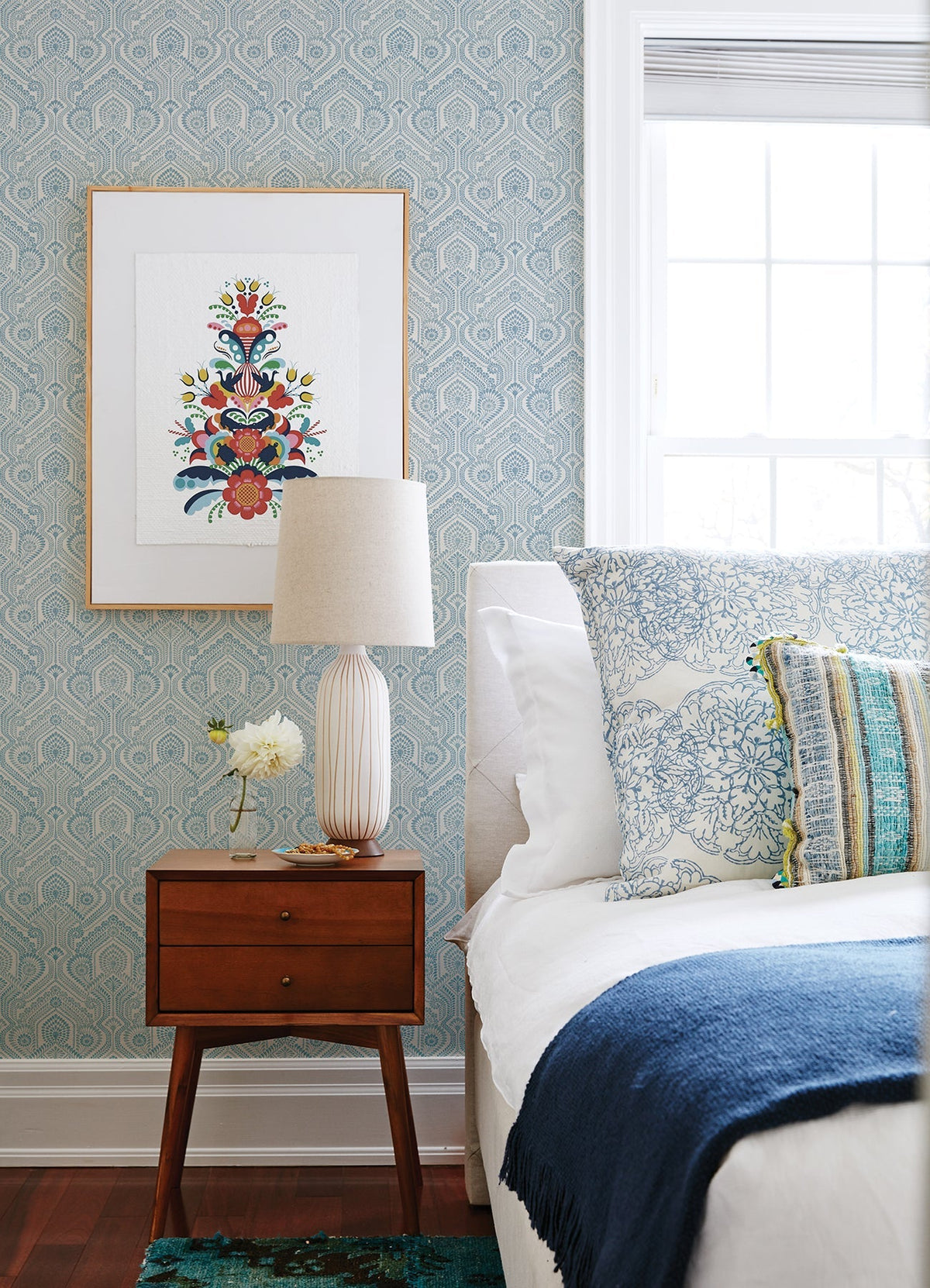 A cozy bedroom featuring a mid-century nightstand, a ceramic lamp, flowers, and a bed adorned with blue and white pillows and blanket. Fernback Ornate Botanical Wallpaper by York Wallcoverings adds pattern behind a framed artwork beside a large window.