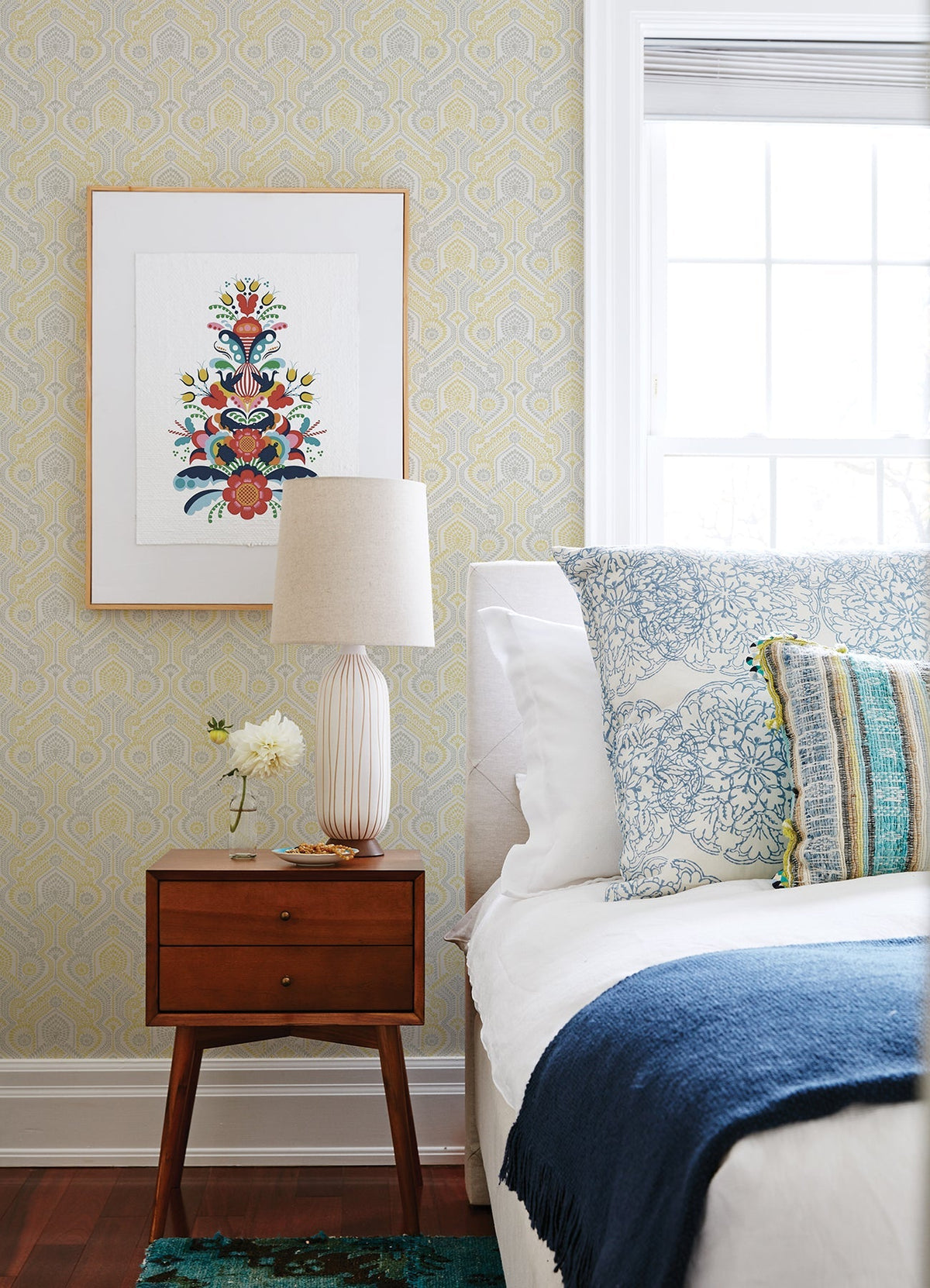A modern bedroom featuring a wooden nightstand, a table lamp, a small flower vase, patterned pillows, a blue throw blanket, and framed art on Fernback Ornate Botanical Wallpaper by York Wallcoverings beside a sunny window.
