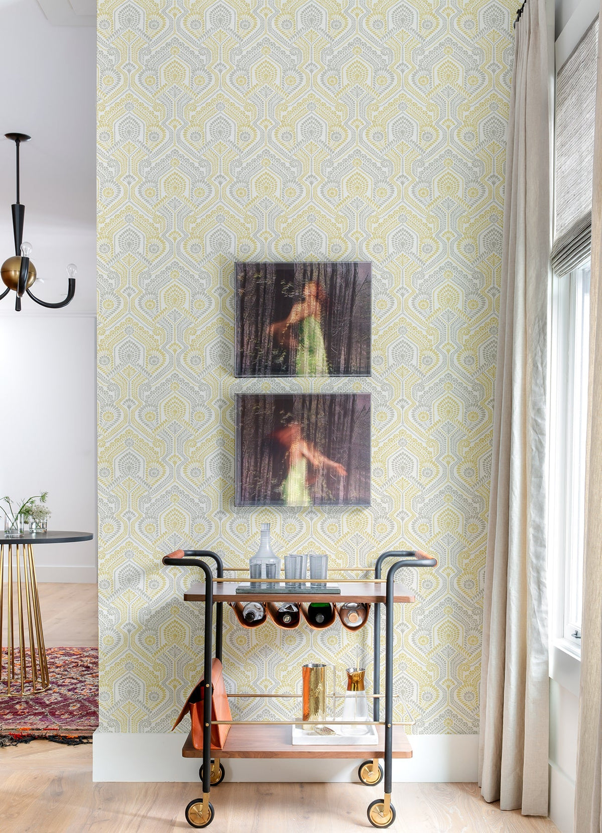 A bar cart with glassware and bottles sits against a wall adorned with Fernback Ornate Botanical Wallpaper by York Wallcoverings, beneath two blurry framed photos of a woman in a green dress. A window with curtains is to the right.