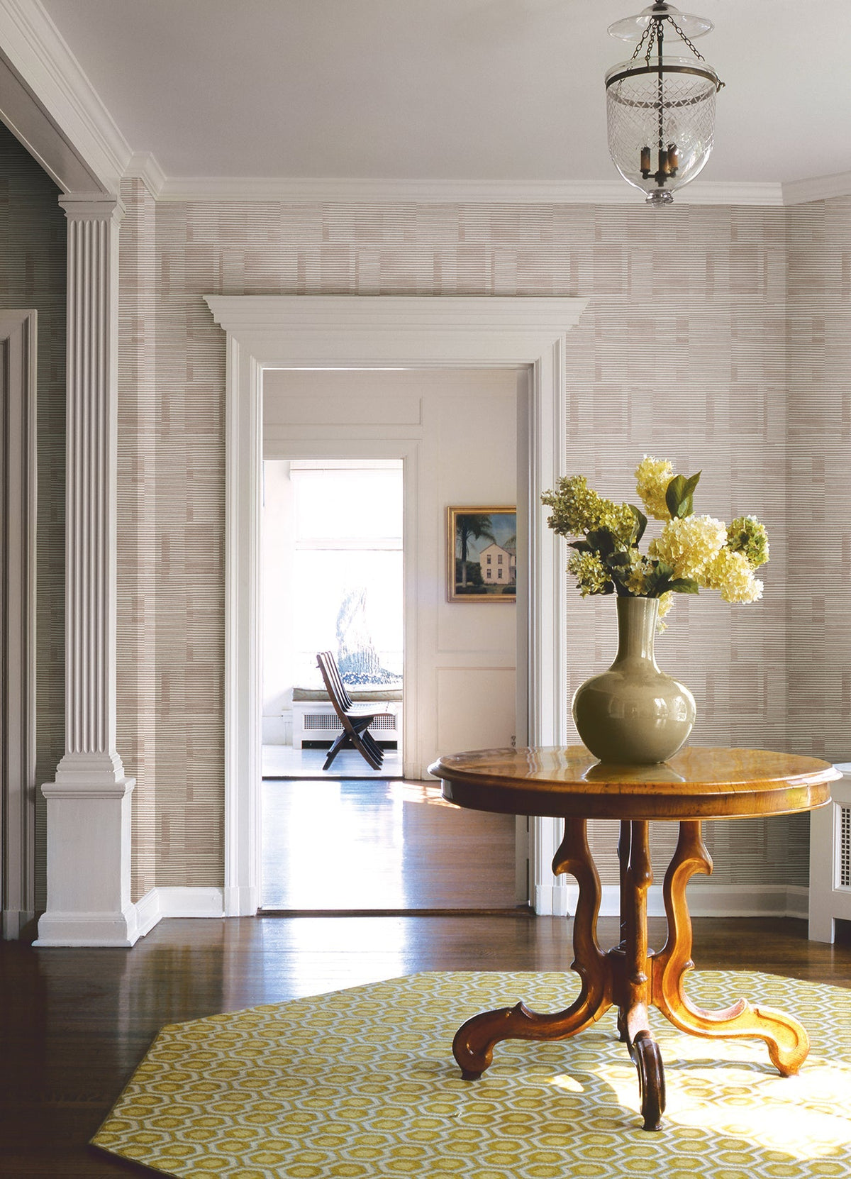 A wooden round table with a green vase of flowers sits on a yellow geometric rug in a hallway with columns, Callaway Woven Stripes Wallpaper by York Wallcoverings, and a hanging light fixture. A sunny room with a chair is visible through the doorway.