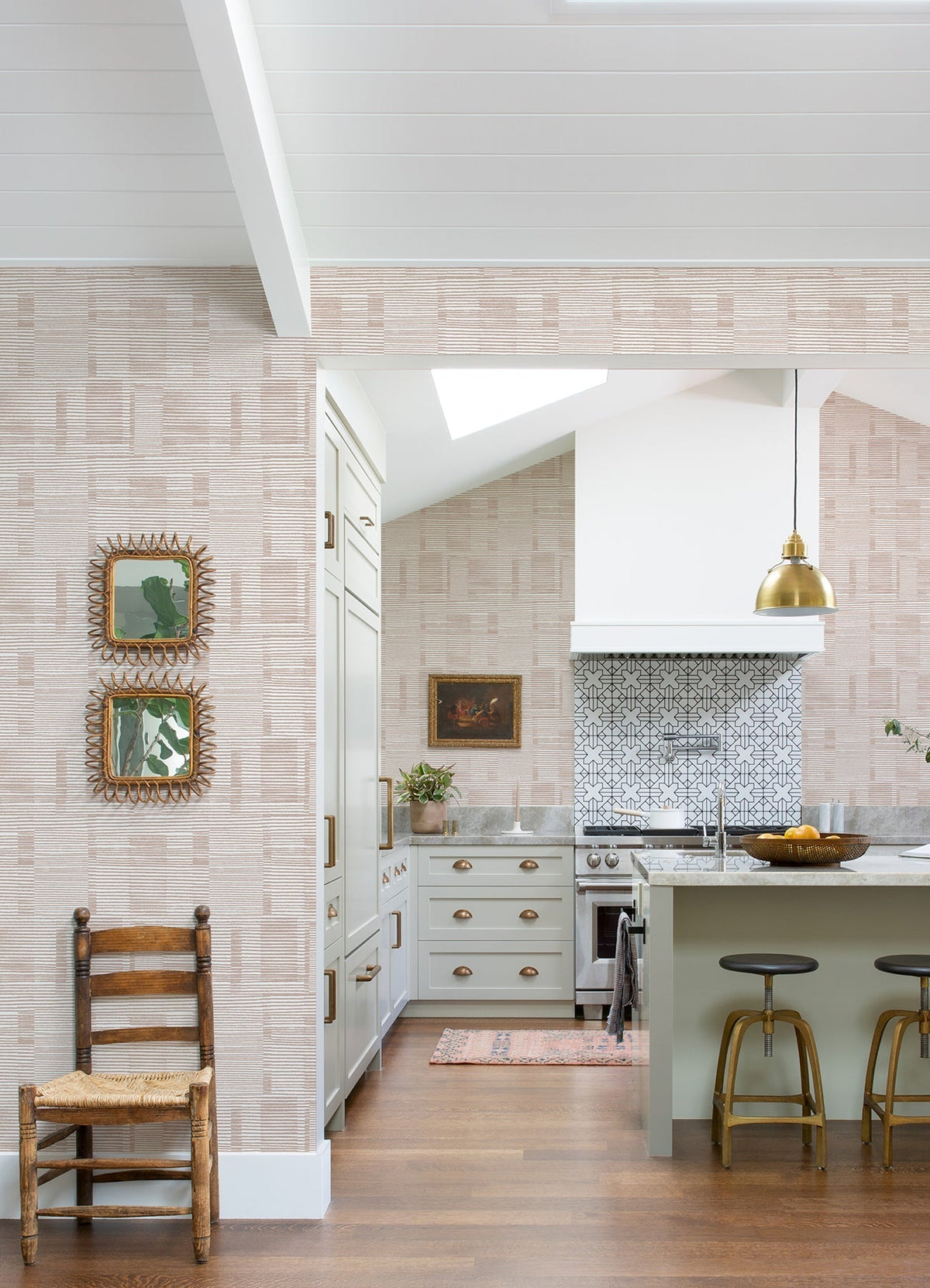 Sunlight fills the kitchen, which features York Wallcoverings’ Callaway Woven Stripes Wallpaper, a green island with three stools, a gold pendant light, and a patterned tile backsplash. A wooden chair sits nearby, while framed art hangs on the wall. Hardwood floors and a vaulted ceiling complete the space.