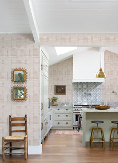 Sunlight fills the kitchen, which features York Wallcoverings’ Callaway Woven Stripes Wallpaper, a green island with three stools, a gold pendant light, and a patterned tile backsplash. A wooden chair sits nearby, while framed art hangs on the wall. Hardwood floors and a vaulted ceiling complete the space.