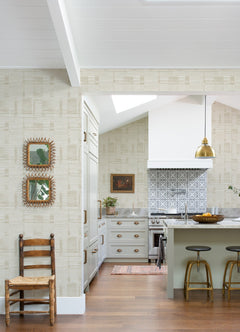 A bright kitchen features light cabinets, a patterned Callaway Woven Stripes Wallpaper by York Wallcoverings as the backsplash, a gold pendant light, two bar stools, decorative mirrors on the wall, and a wooden chair on a hardwood floor.