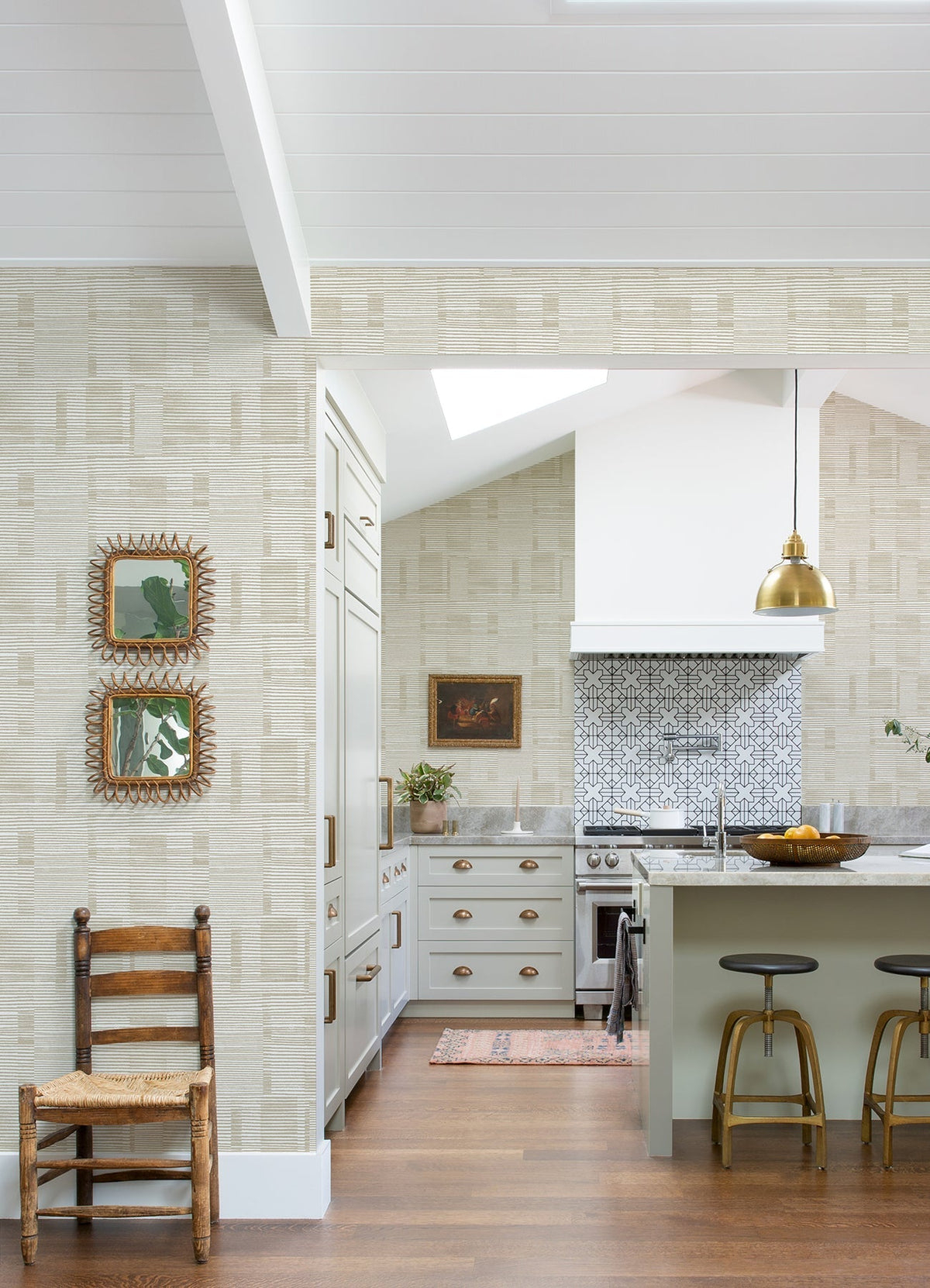 Bright kitchen featuring a patterned backsplash, white cabinets with gold hardware, and a green island accompanied by two stools. A wooden chair sits against the wall adorned in Callaway Woven Stripes Wallpaper from York Wallcoverings, where framed mirrors also hang. Above the island is a gold pendant light.