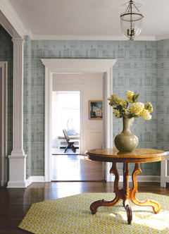 A round wooden table sits on a yellow rug in a bright entryway adorned with Callaway Woven Stripes Wallpaper by York Wallcoverings, featuring a vase with yellow flowers and columns framing a doorway that leads to a sunlit room in the background.
