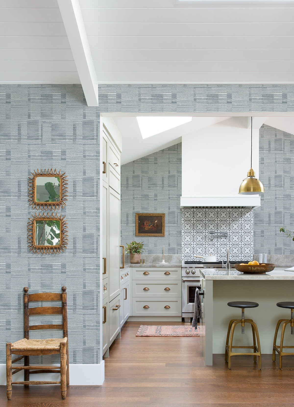 A modern kitchen features Callaway Woven Stripes Wallpaper by York Wallcoverings, a white stove, gold pendant light, three barstools, and wooden accents. Two framed mirrors and a wooden chair are set against the wall in the foreground.