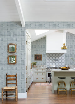 Bright kitchen featuring York Wallcoverings’ Callaway Woven Stripes Wallpaper, gold pendant lights, barstools at a green island, a wooden chair, and two framed mirrors on the wall. White cabinets, a patterned backsplash, and wooden floors complete the cozy space.