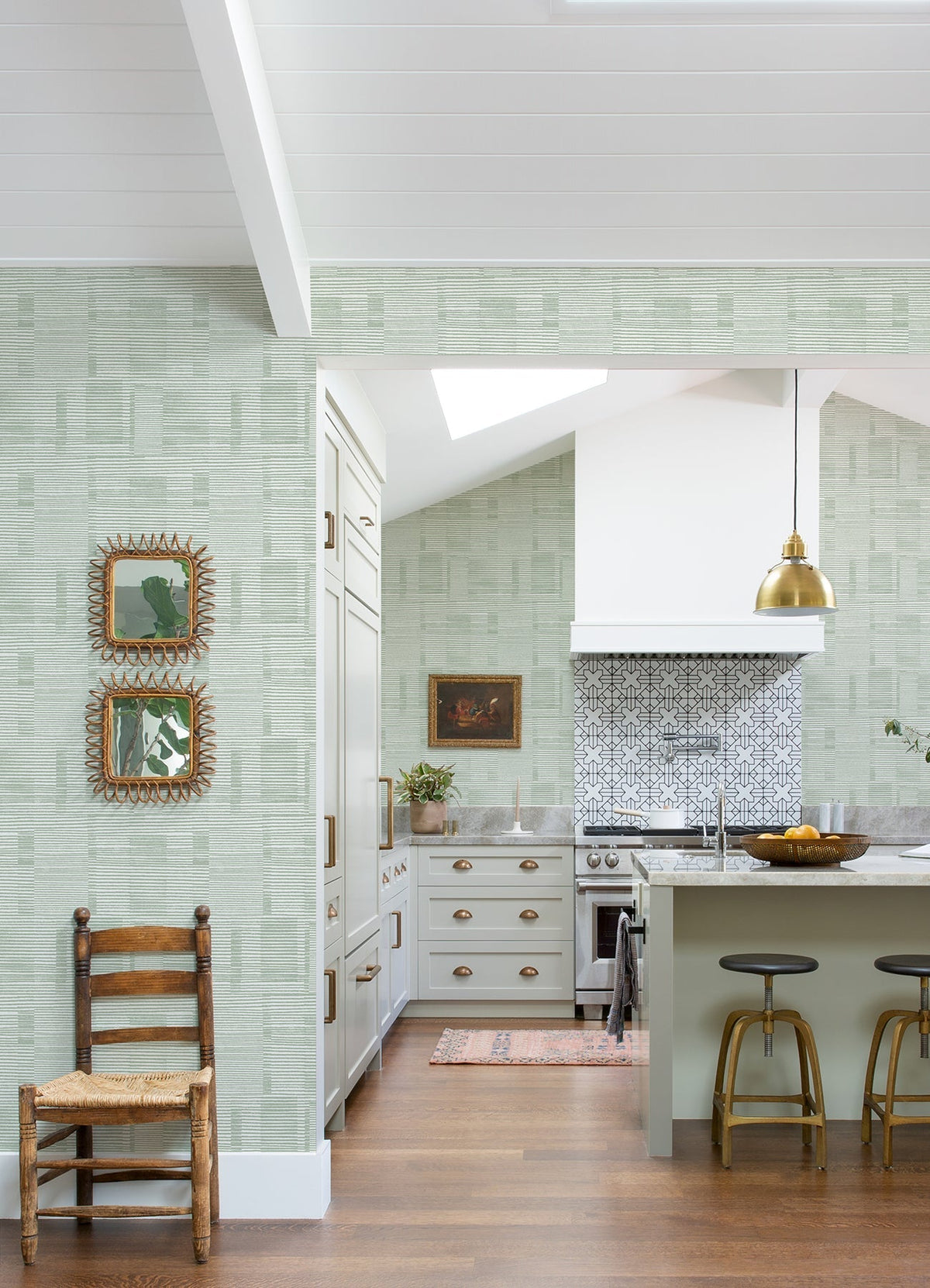 The kitchen features airy, high white ceilings and wood floors, with a bright atmosphere. The walls are adorned with Callaway Woven Stripes Wallpaper by York Wallcoverings in green patterns. The space includes a wooden chair, two mirrors, gold light fixtures, a white stove, an island with stools, and a patterned backsplash.