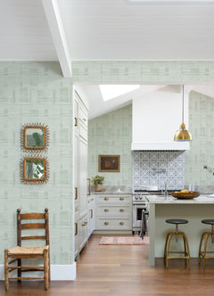 Bright kitchen featuring Callaway Woven Stripes Wallpaper by York Wallcoverings in green, complemented by white cabinets, a gold pendant light, patterned backsplash, wooden stools, a rustic chair, and two framed mirrors on the wall. Natural light fills the space.