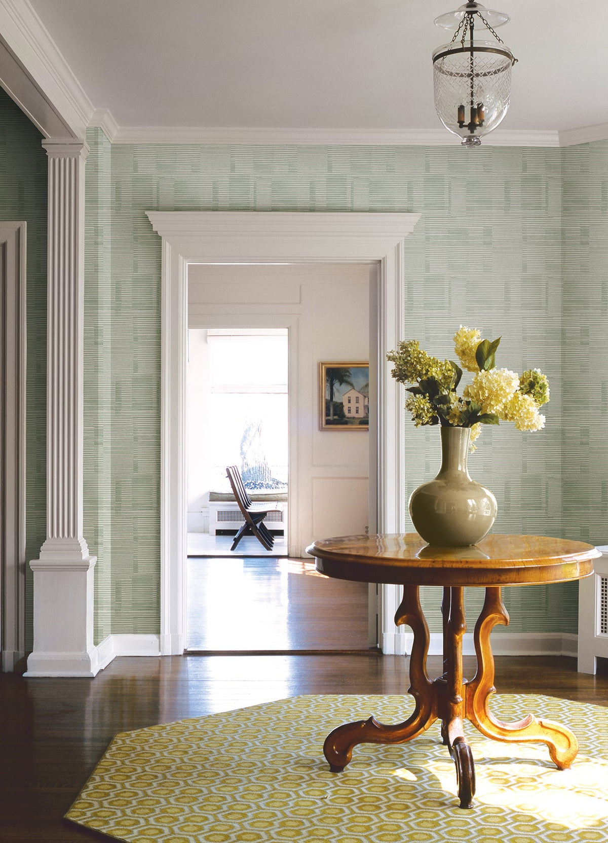 A round wooden table with a vase of yellow flowers sits on a yellow patterned rug in a bright hallway decorated with York Wallcoverings’ Callaway Woven Stripes Wallpaper, complemented by white trim, columns, and a hanging glass light fixture.
