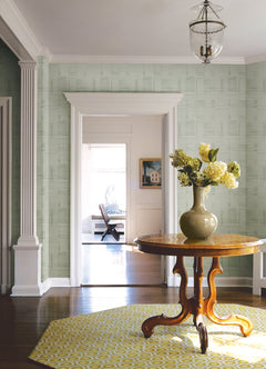 A round wooden table with a vase of yellow flowers sits on a yellow patterned rug in a bright hallway decorated with York Wallcoverings’ Callaway Woven Stripes Wallpaper, complemented by white trim, columns, and a hanging glass light fixture.