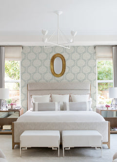 A bright, elegant bedroom featuring a large bed with two benches at the foot, symmetrical nightstands topped with modern lamps, and two windows. The walls are adorned with Zaria Topiary Wallpaper by York Wallcoverings, and an oval gold-framed mirror hangs above the headboard.