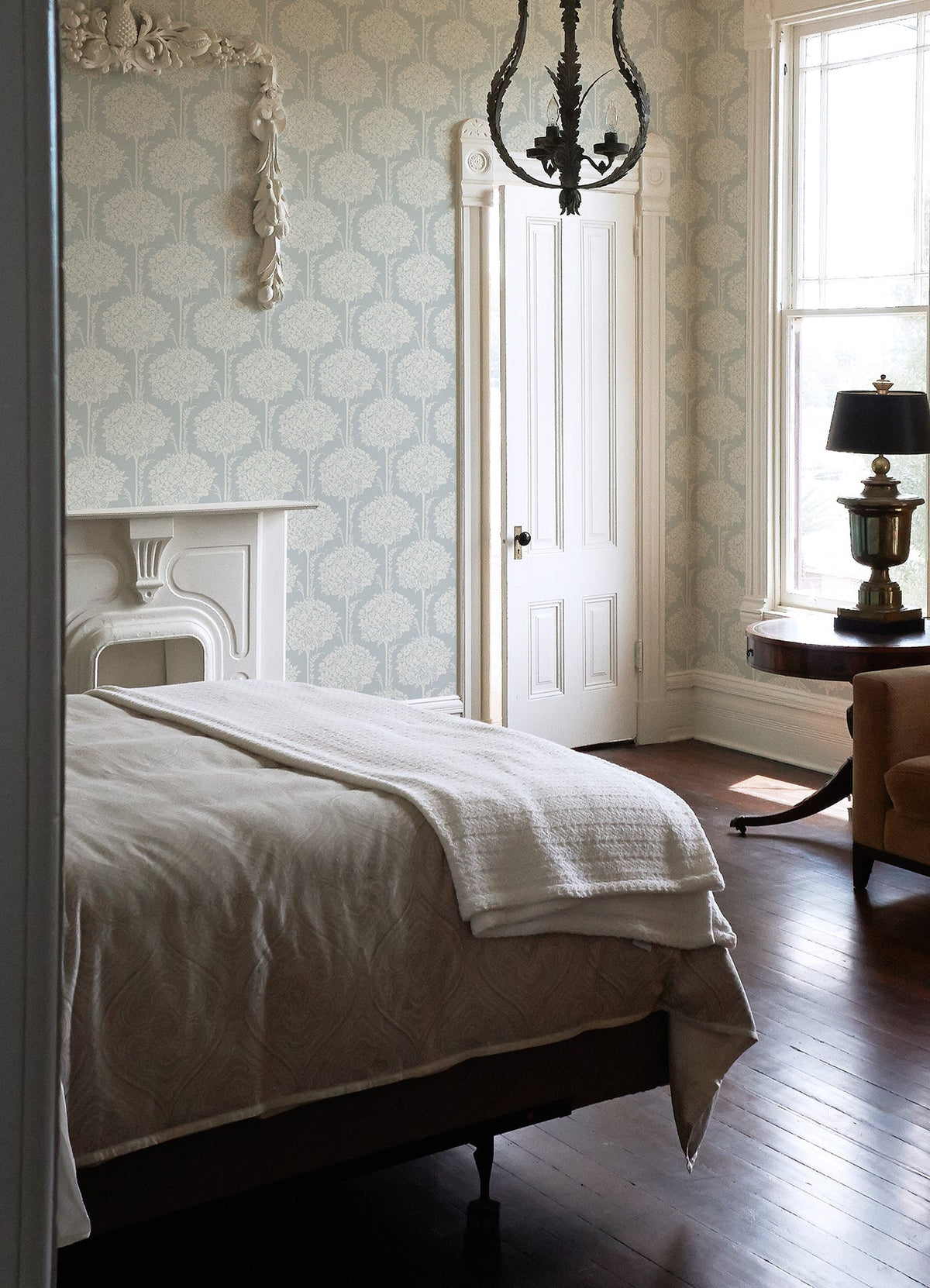 A cozy bedroom featuring a bed with white bedding, an ornate fireplace, chandelier, round table with lamp, armchair, and a large window letting in natural light is beautifully complemented by York Wallcoverings’ Zaria Topiary Wallpaper.